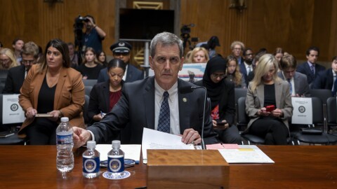 A man sits a huge wooden desk with three bottles of water and pages that he turns through in a binder on the table. People are seated behind him. 