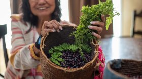 An older woman, wearing glasses, holds a basket of huckleberries while seated at a kitchen table.