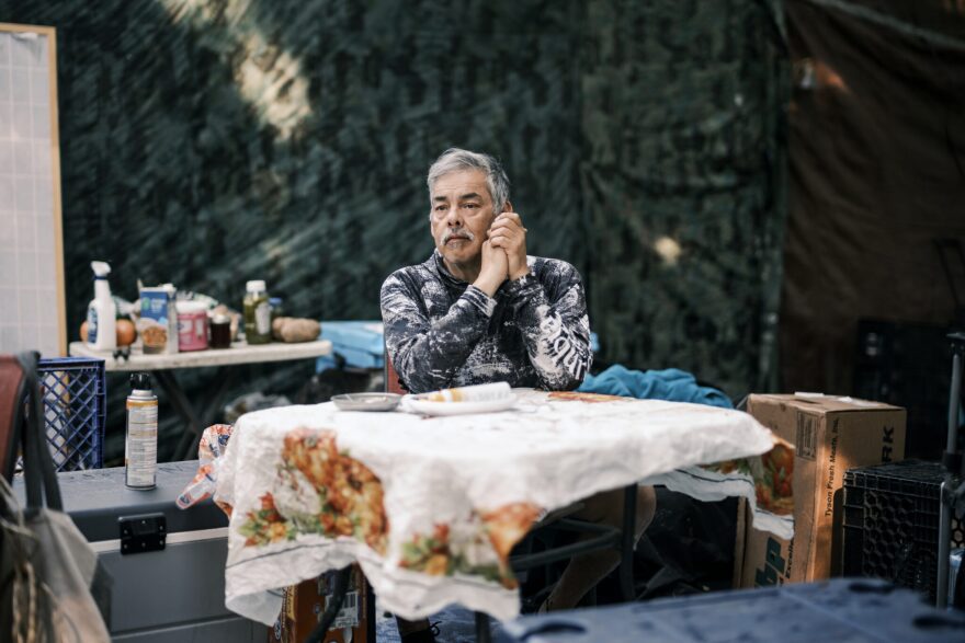 An older man sits at a table wearing a white and black hoodie with his hands clasped together.