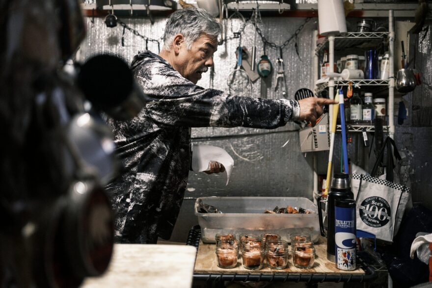 A man wearing a white and black hoodie with graying hair stands in front of a container full of chopped-up salmon pieces and jars full of salmon. Off to the side is a wall with hanging cutlery.