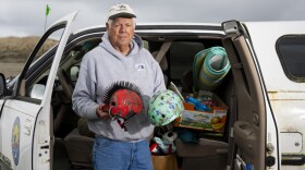 An older man stands in front of the open cab of an older white pickup trucked filled with all sorts of items as he holds two bicycle.