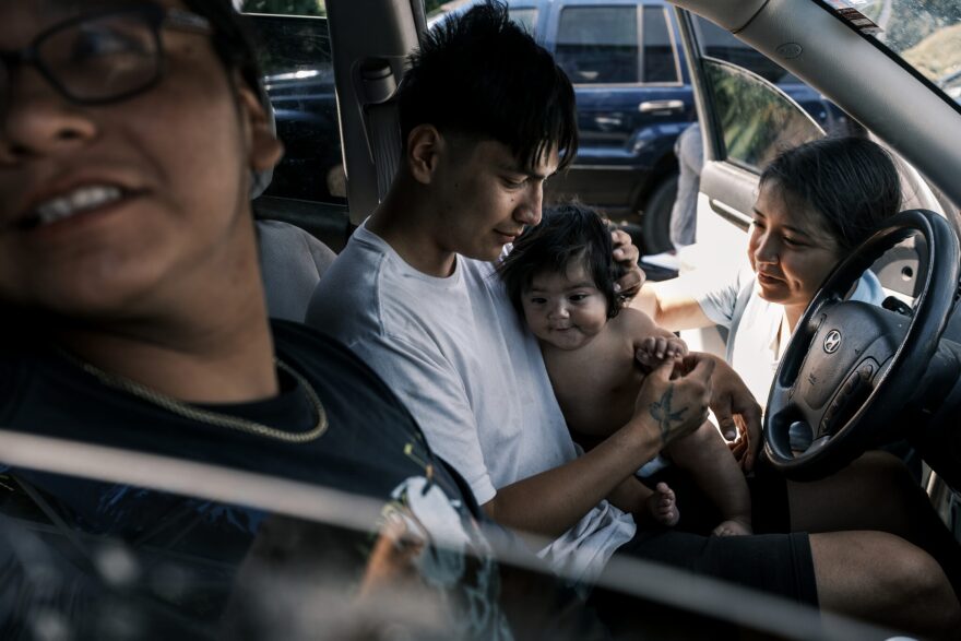 A young man sits in the front seat of a car holding a baby, while a young woman leans in through the car door to rub the baby's head. Another person on the passage side looks out the window. 