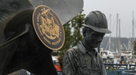 A statue of a worker near the water front in Bremerton, next to a round plaque commemorating the 100th anniversary of the Puget Sound Naval Shipyard