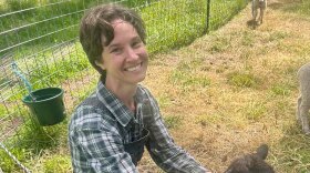 A woman smiles while wearing overalls and a plaid shirt crouched down while petting sheep. 