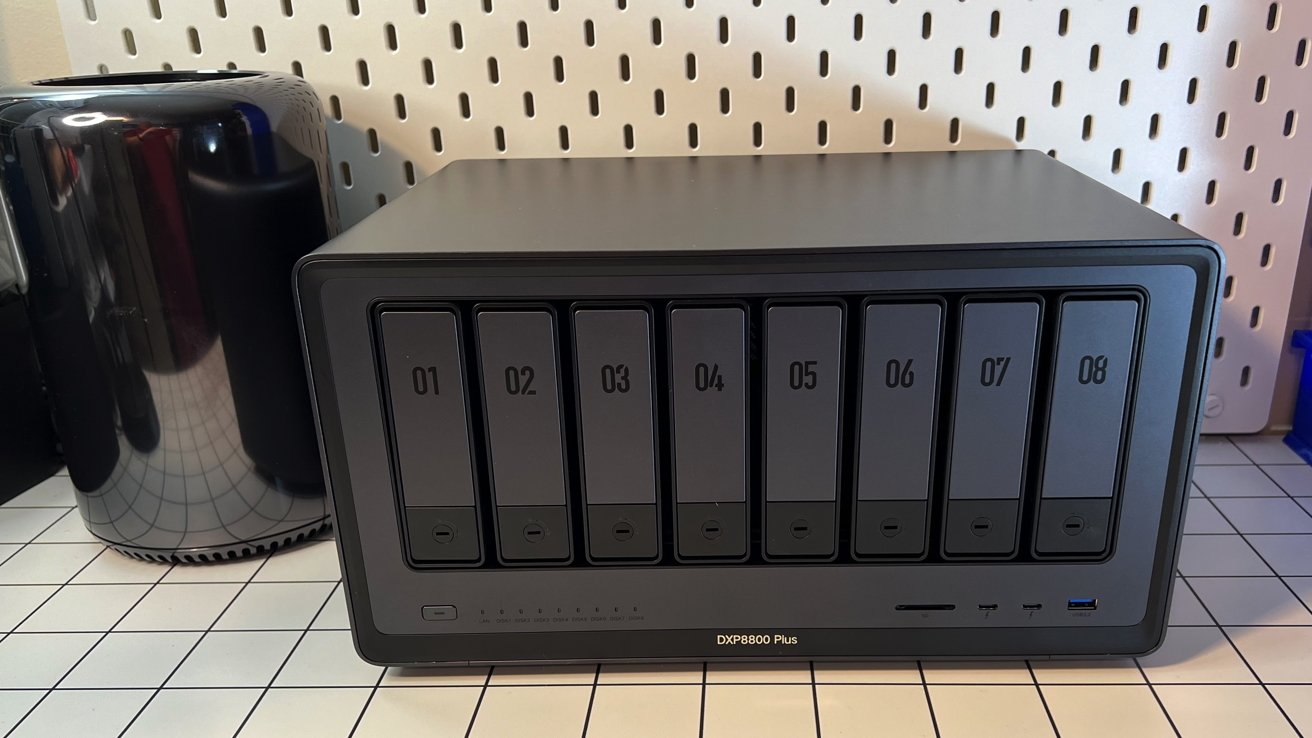 A black desktop NAS device with eight numbered hard drive bays on a white grid table with a reflective black Mac Pro in the background.