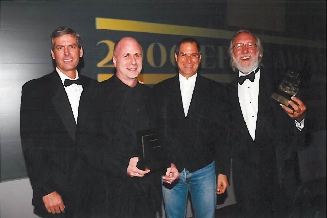 Four men standing together, three in tuxedos and one in jeans and a blazer, holding trophies and smiling.