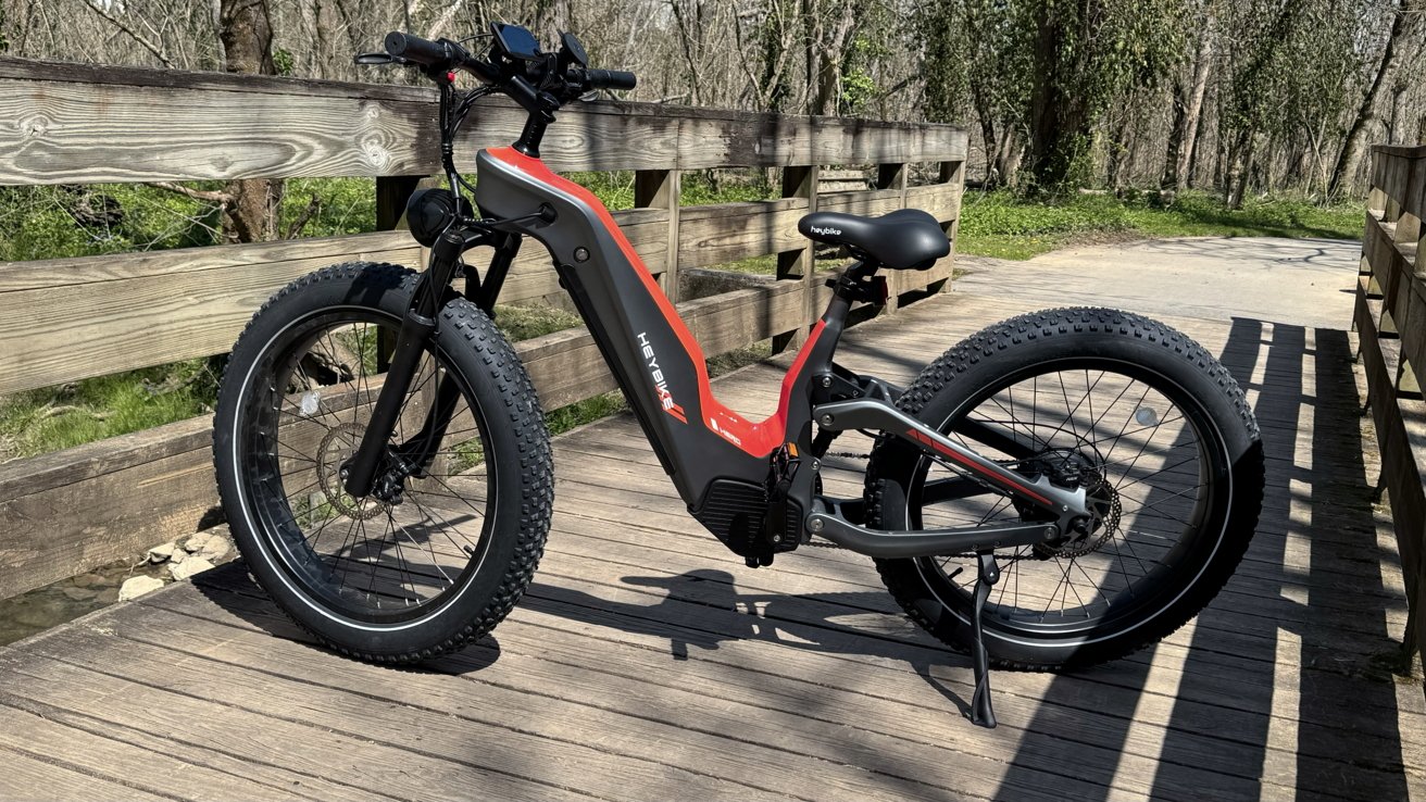 Heybike Hero on a small walking bridge with a path going by trees in the background. The bike is gray with orange accents.