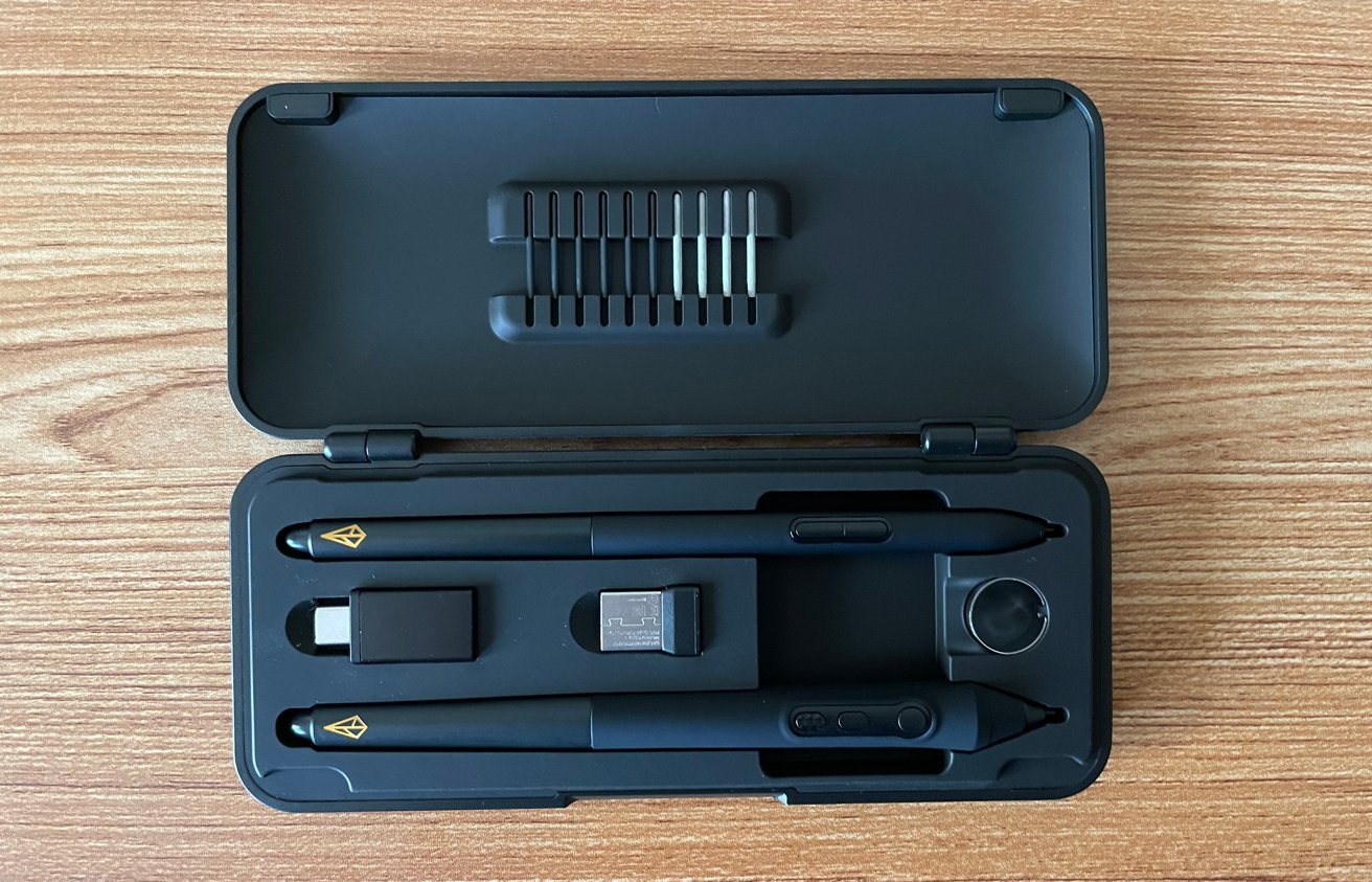 Opened black case on a wooden surface containing pens, small electronic accessories, and spare pen nibs in a holder.