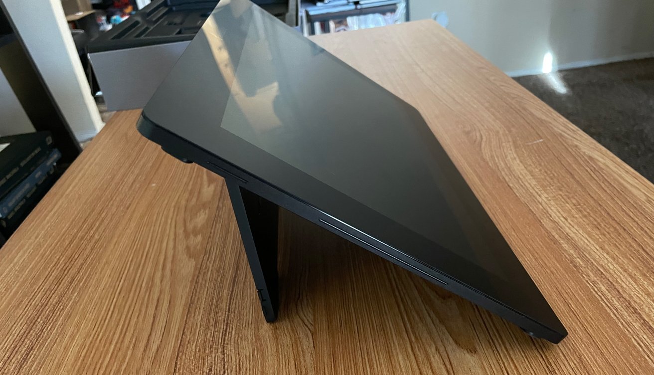 Black electronic device with a stand, angled on a wood-grain table, surrounded by office items in the background.