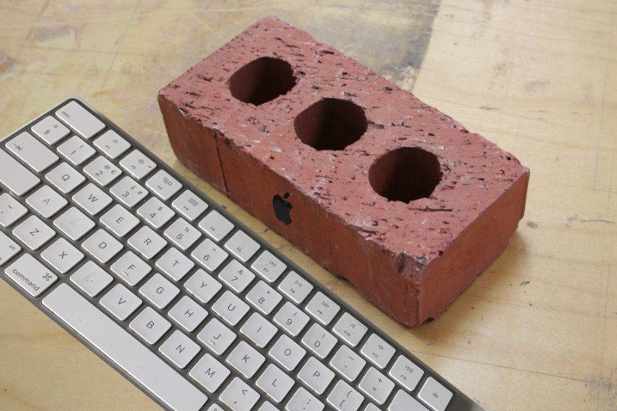 Red brick with three holes placed next to a white computer keyboard on a wooden surface.