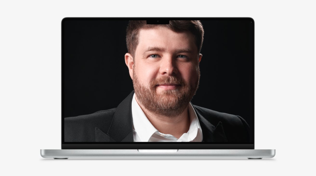 Headshot of a bearded man in formal attire displayed on a laptop screen with a dark background.