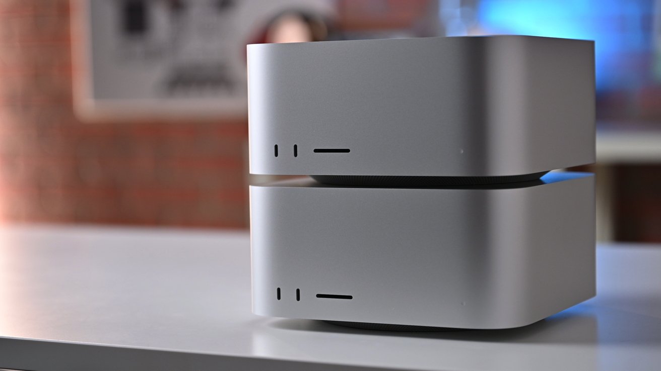 Two stacked, silver Mac Studios, sitting on a white surface. Blurred background features a brick wall and a sign.