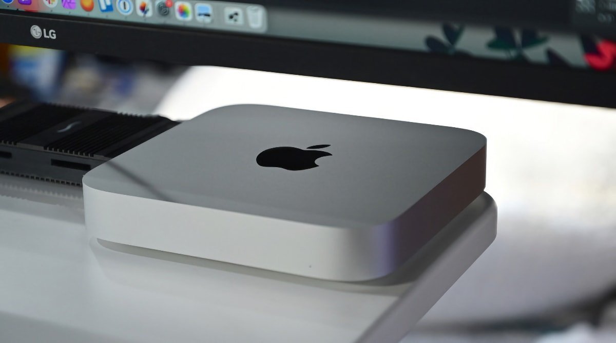 A silver Apple Mac Mini computer on a desk, with an LG monitor in the background displaying a macOS desktop.