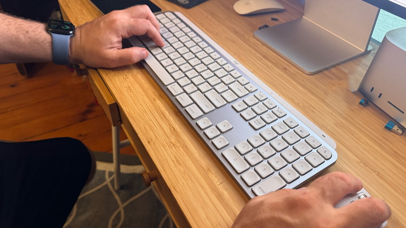 Hands typing on a silver keyboard on a wooden desk, with a wristwatch visible on the left wrist.