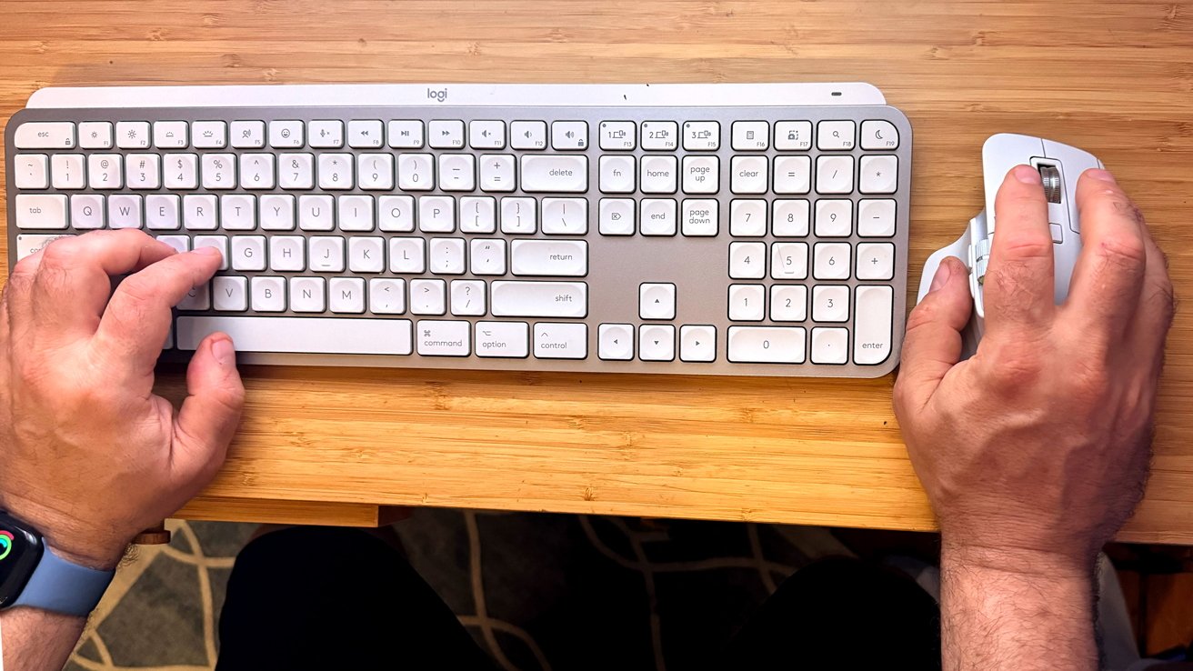 Hands typing on a white keyboard with a number pad while using a computer mouse on a wooden tabletop.