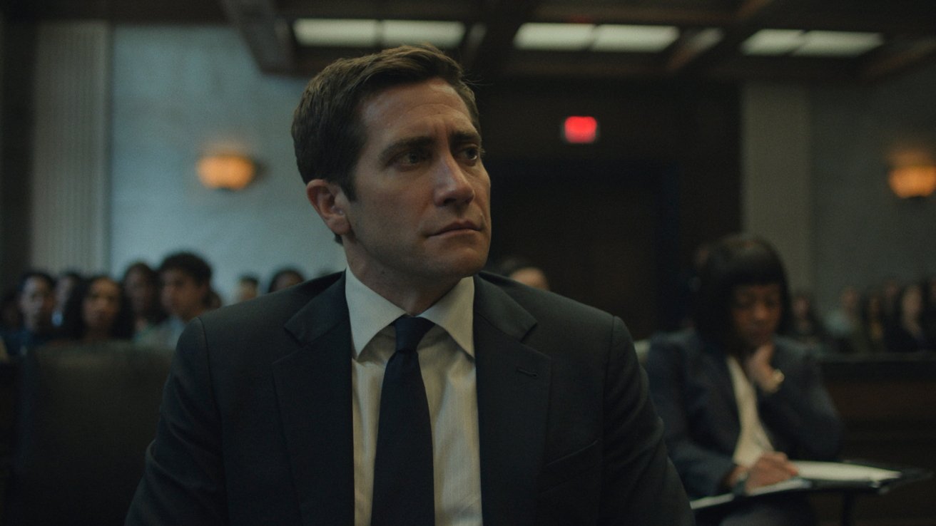 Man in a suit and tie sits in a courtroom, appearing serious, while a woman writes in a notebook in the background.