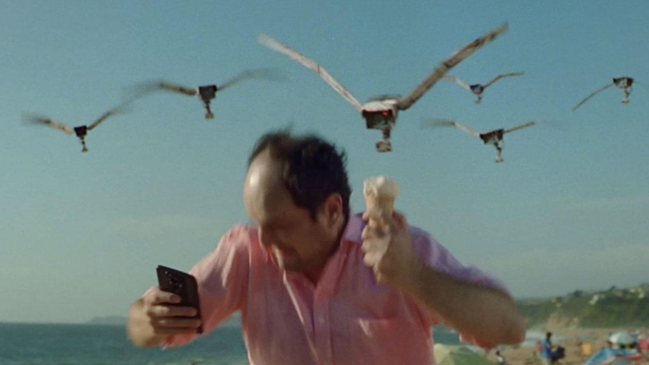 Man dodging robotic birds while holding an ice cream and a phone on a beach.