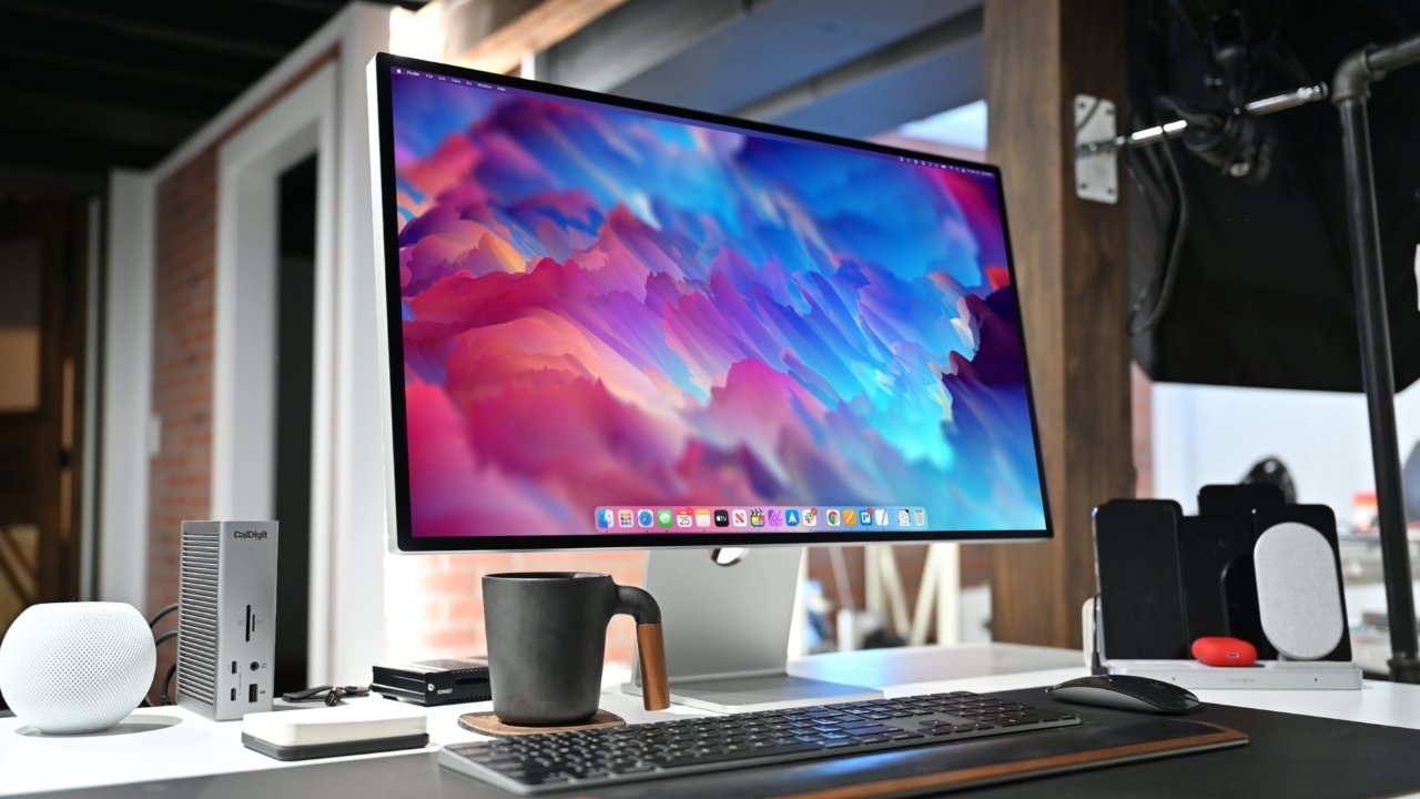 Modern workspace with an Apple Studio Display showing colorful abstract art, a keyboard, mouse, black mug, smart speaker, and various electronic devices on a white desk.