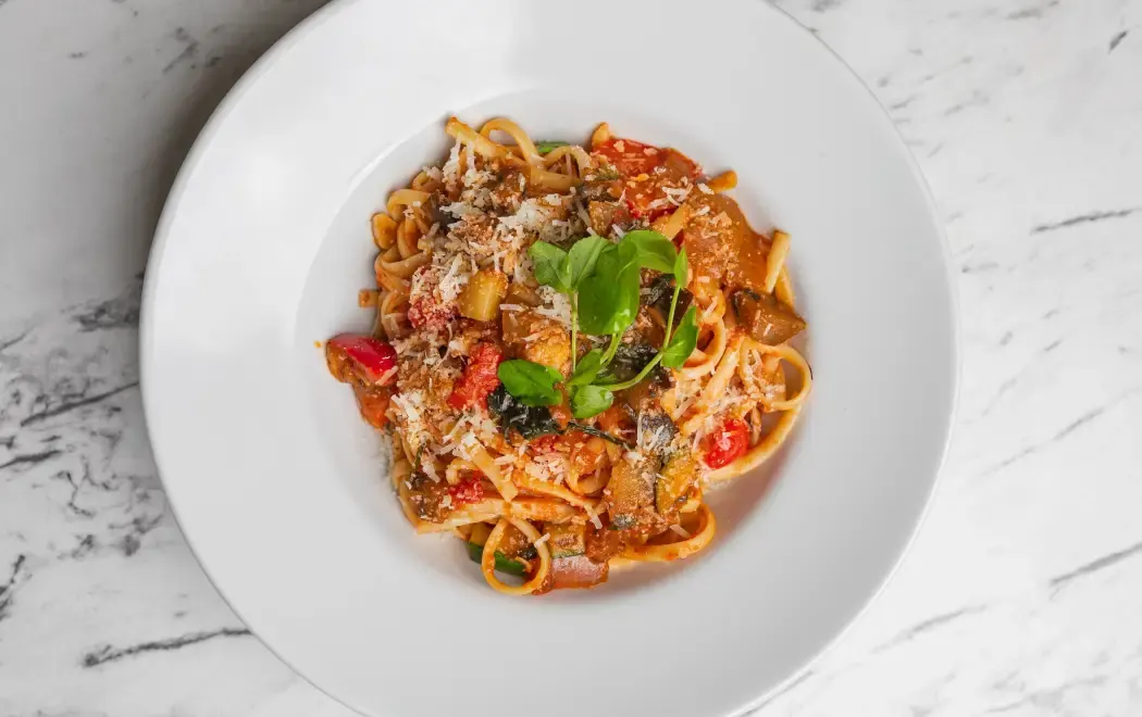 A white plate of pasta topped with grated cheese, vegetables, and basil, set on a marble surface.