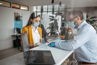 Bank teller discussing paperwork with customer at bank counter wearing protective gloves and face mask. Office with acrylic glass partition on desk. Acrylic glass wall - protection against coughs and spitting, protection against viruses.