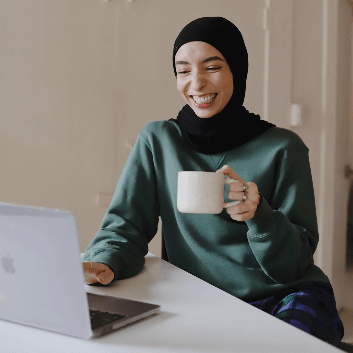 This is a picture of a woman sat down behind a laptop, drinking a hot drink