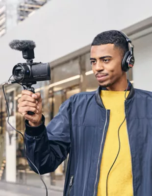 A man wearing headphones holds a Sony camera with a shooting grip and microphone attached. 