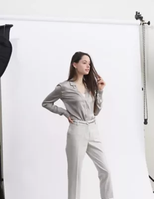 A woman in a light grey outfit poses in front of a white backdrop in a photo studio.