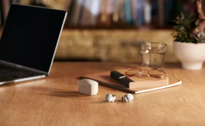 White wireless earbuds sit next to their charging case on a wooden table