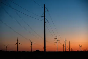 Power lines, seen in twilight, extending from the foreground to the horizon, with wind turbines on either side. The sky is deep red along the horizon.