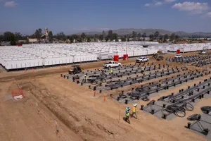 An aerial view of dozens of large battery units lined up in rows. They resemble shipping containers. In the foreground, a few pickup trucks. The sky is clear and blue.