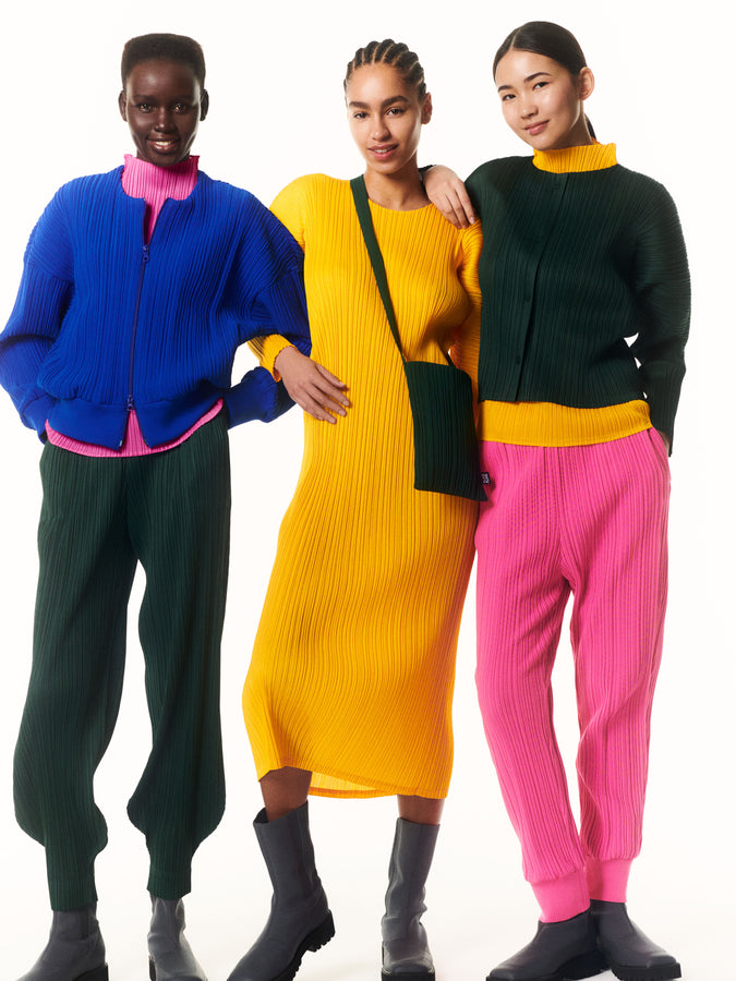 Three models wearing colourful pleated clothing posing in front of white background.