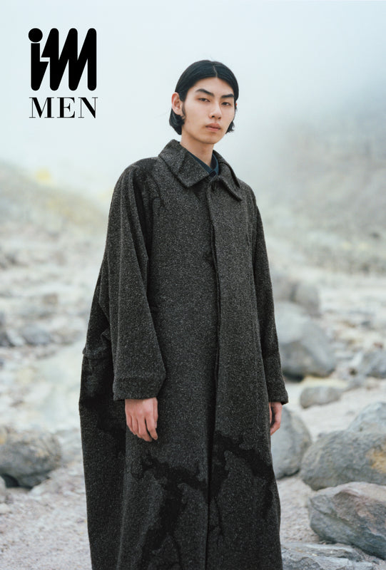 Model wearing long charcoal grey coat in rocky landscape covered in mist. Top left corner: IM MEN logo in black on white background. 