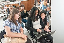 Trzy młode kobiety siedzą na krzesłach; siedząca pośrodku na kolanach trzyma laptop, jej sąsiadka z lewej strony kadru patrzy na ekran komputera, ta z prawej strony sięga w kierunku klawiatury.