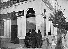 Photo[permanent dead link] of large white building with one signs saying "Moritz Schiller" and another in Arabic; in front is a cluster of people looking at poster on the wall.