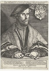 A middle aged man, Wilhelm, Duke of Jülich-Cleves-Berg, is sitting at a table. He is dressed in a soft cap that falls to the side of his head. He is wearing fur-trimmed robes, and nestled in the edges of the robes is a chain, with a cross at its nadir. His hands are folded on the table before him. He is wearing several rings, and one hand holds a pair of gloves. The family crest hangs on the chair behind him.