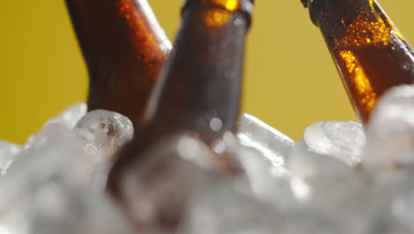 close up of glass bottles of cold beer or soft drinks chilling in ice filled bucket against yellow background