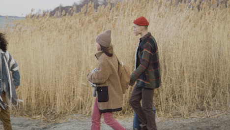 side view of a teenage boy and girl approaching to two friends wearing winter clothes walking in a wheat field near of sea on a cloudy day