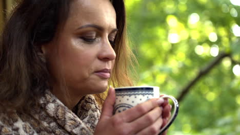 close up fhd shot of a brunette middle-aged woman smelling freshly poured tea in a mug she holds with both her hands with blurred trees in the background