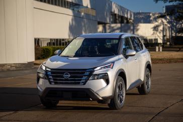 A front left 3/4 view of a 2024 Nissan Rogue parked on cement in front of a white building.