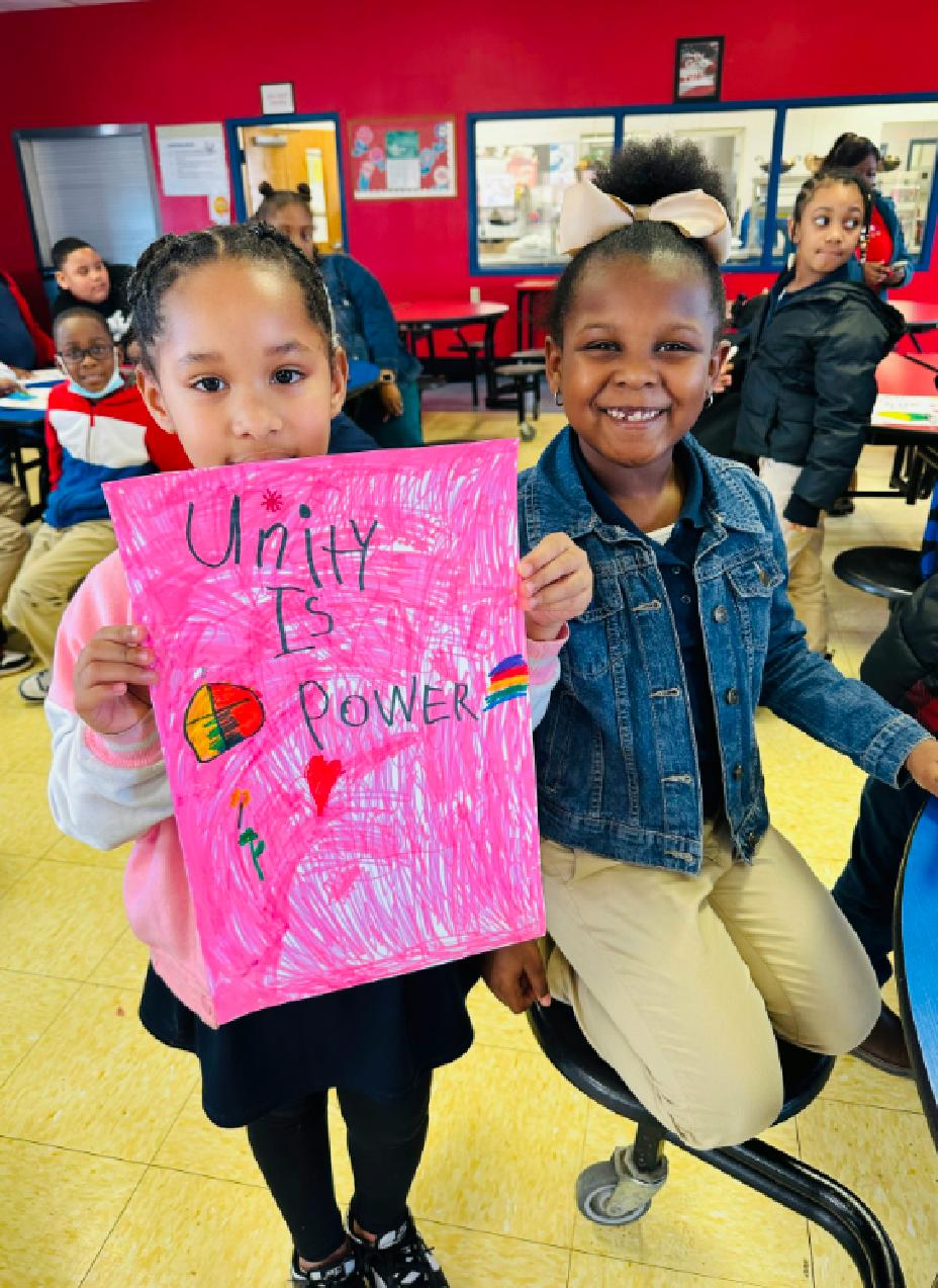 In a classroom, a young girl holds a hand drawn poster that reads “Unity is Power” while standing next to another smiling girl.
