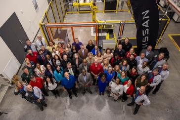 A class of Nissan workers and instructors standing in a warehouse all smiling towards the camera that is overhead.