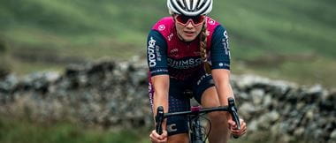 Cyclist Dr Sophie Earl riding towards the camera with a stone wall and green field in the background