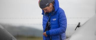 Cyclist Rebecca Richardson zipping up her blue coat while listening to music on earbuds with grey sky in the background