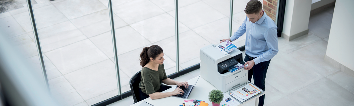 Man sat at desk with laptop and DCP-L3550CDW in background