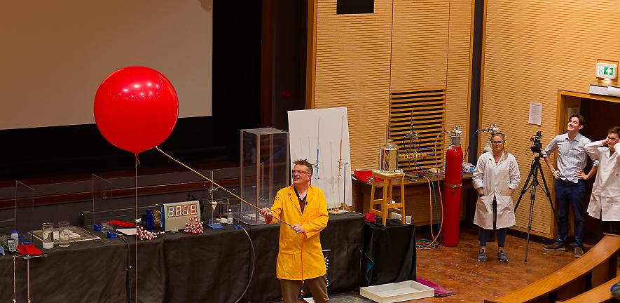Peter Wothers on stage in goggles & lab coat about to light a hydrogen-filled red balloon
