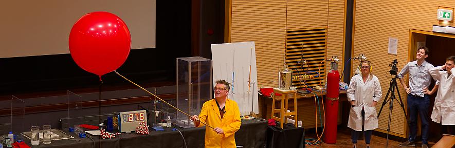 Peter Wothers on stage in goggles & lab coat about to light a hydrogen-filled red balloon