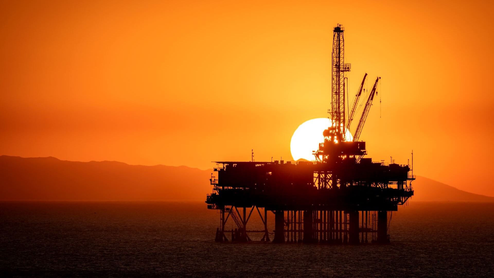 Silhouette of offshore oil platform set against orange sky and setting sun