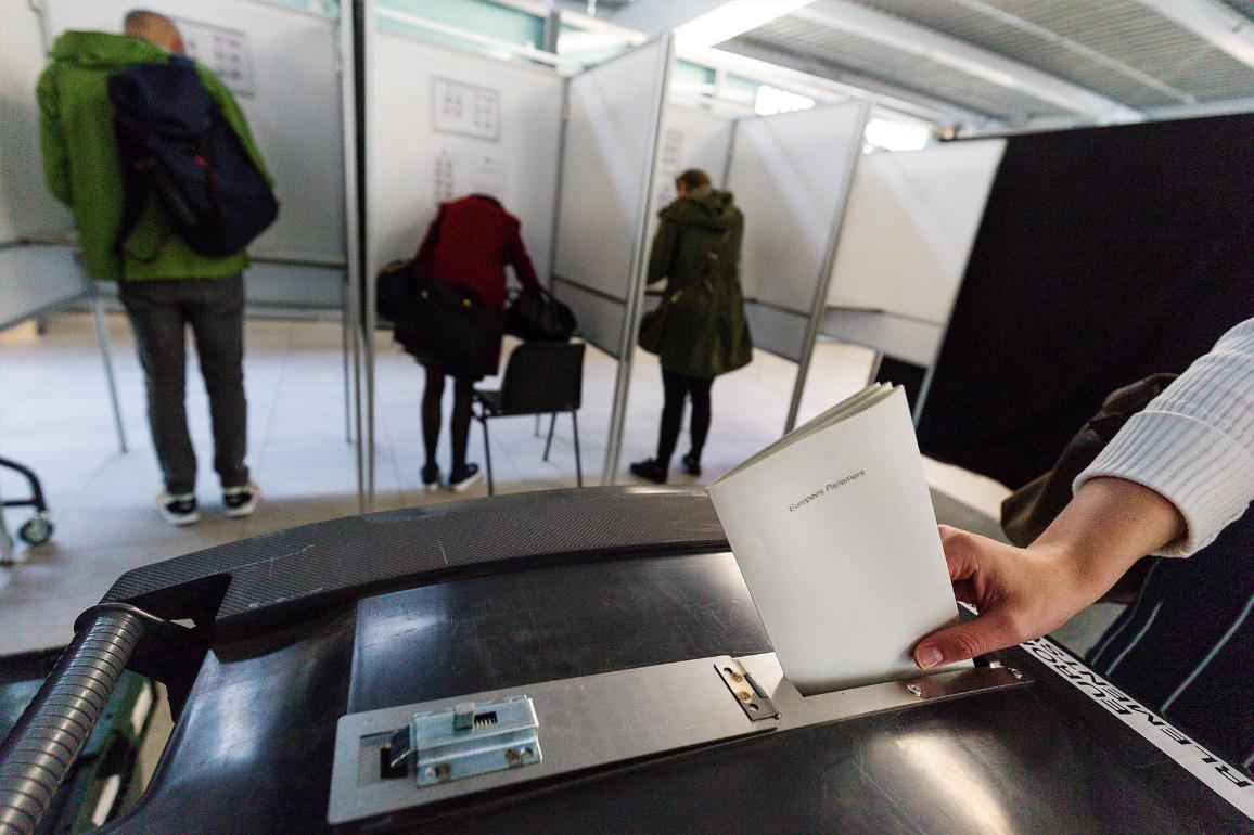 A hand putting a paper in a ballot box. We see voting booth in the background.