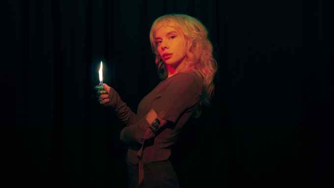 Pixey, with bleach-blond hair and wearing a brown shirt with a buckle attached to sleeves, stands against a black background holding a lighter with a flame