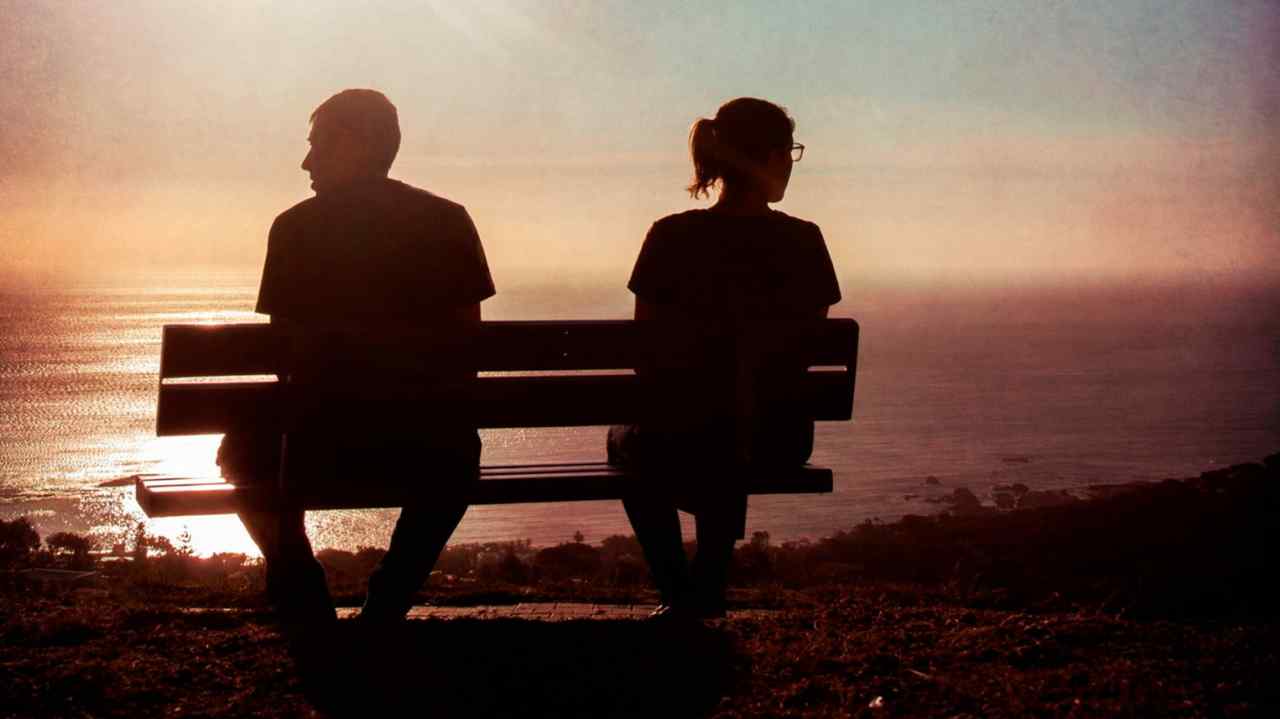 A young couple, seen from behind, sit on a bench overlooking the sea at sunset. They are looking away from each other